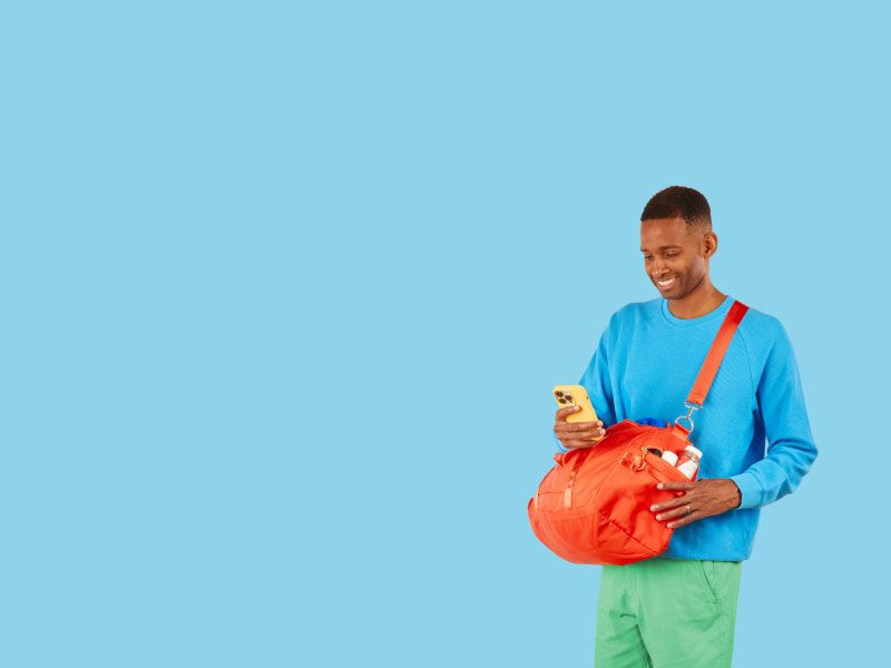 A man smiling and looking at his mobile phone while wearing a bright blue shirt and green shorts with an orange travel bag slung over his shoulder in front of a light blue background