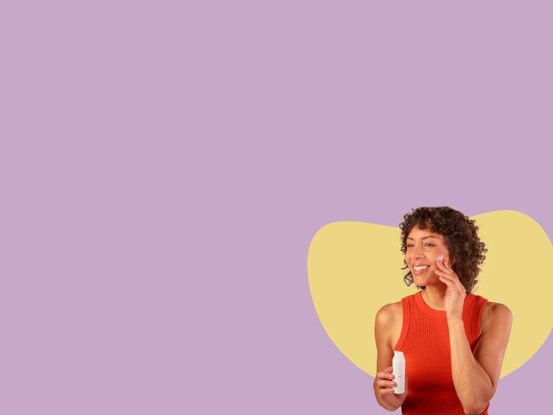 A smiling woman wearing a red tank top is applying Topical Spironolactone cream to her face
