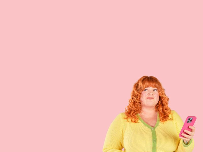 A woman with red hair is wearing a yellow and green sweater while holding a pink phone and looking confused