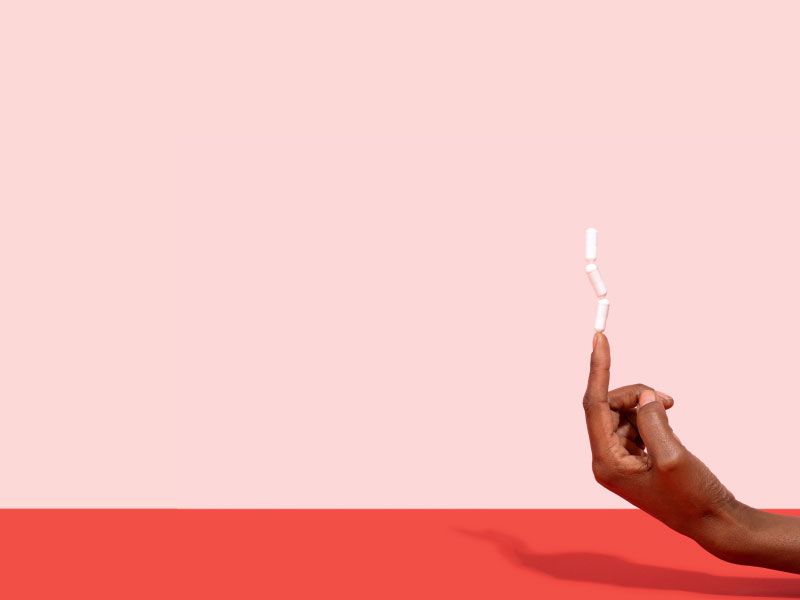 A woman's hand with 3 Boric Acid capsules balancing on her pointer finger on a red surface with a pink background