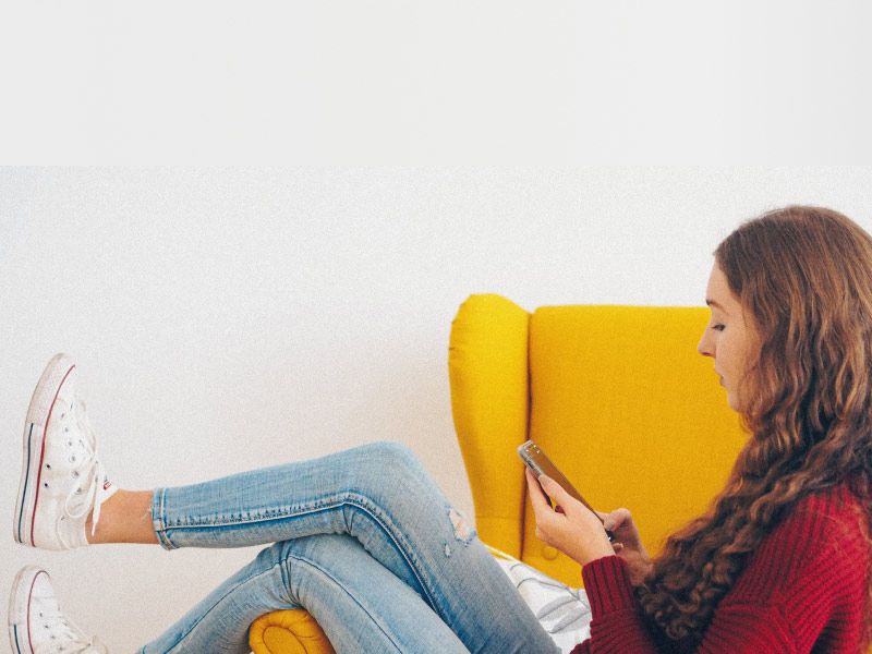 A female with brown hair wearing a red sweater, blue jeans and white shoes is sitting on a yellow chair looking up information on a mobile phone