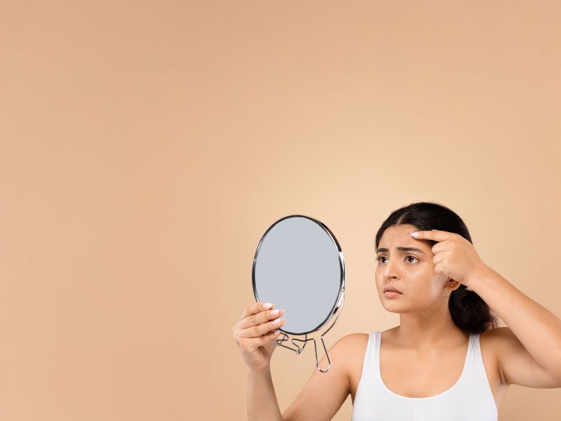 A woman wearing a white tank top is looking into the mirror while touching her forehead