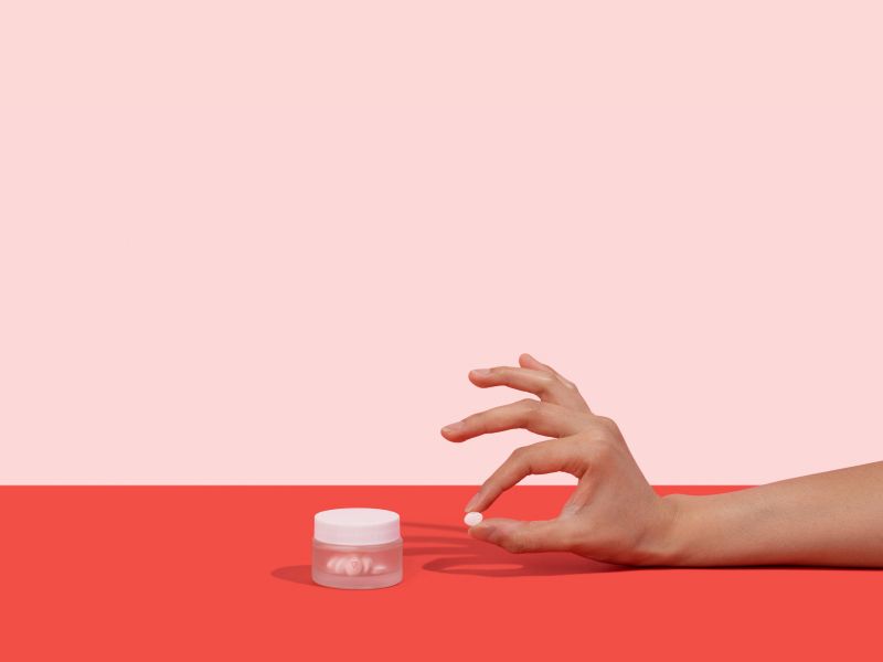 A woman's hand holding a Fluconazole pill next to a Wisp glass jar on a pink and red background