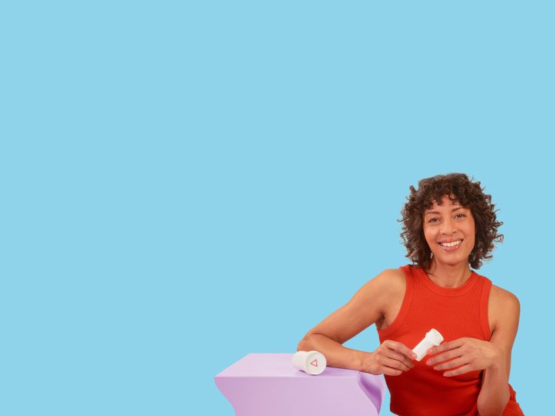 A woman wearing a red tank top is smiling while holding a Wisp pill vial next to a purple table