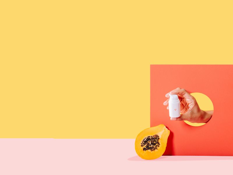 A person's hand holding a Wisp medication bottle through an orange cardboard cutout next to a cut open papaya with a yellow and pink background