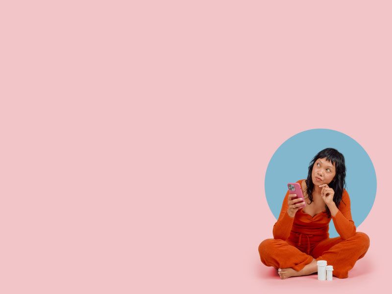A woman wearing a red shirt and pants its sitting on a pink surface with a mobile phone and a confused look on her face
