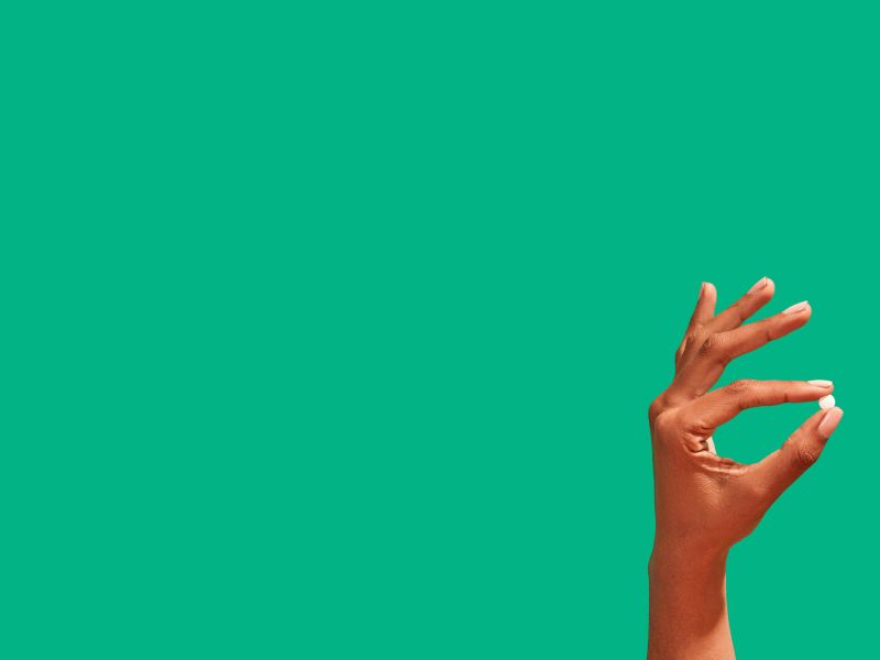 A woman's hand holding an emergency contraception pill in front of a green background