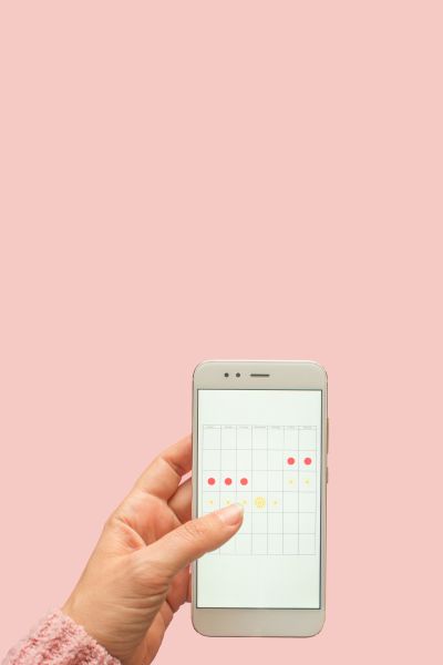 A woman's hand holding a mobile phone with a calendar tracking her menstrual cycle with tampons and menstruation medication on a pink surface in the background