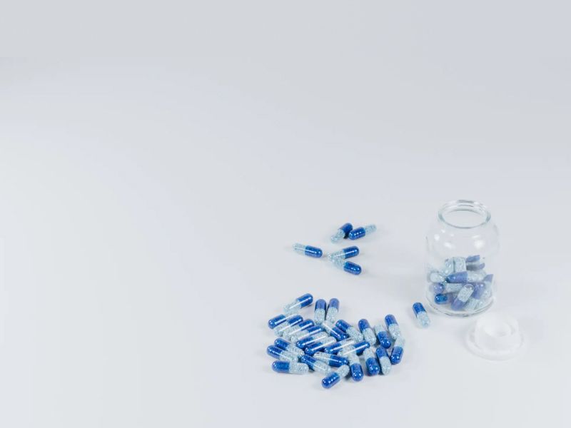 A clear pill bottle with blue and white pills inside and scattered around the bottle