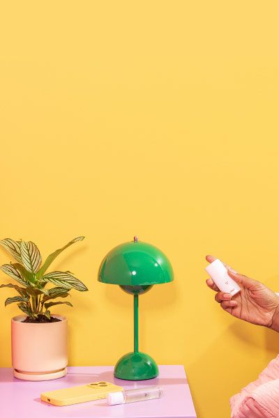 A woman in bed holding Wisp OMG Cream next to a purple nightstand with a green lam, a plant, Wisp Harmonizing Lube, and a mobile phone on it with red and yellow walls
