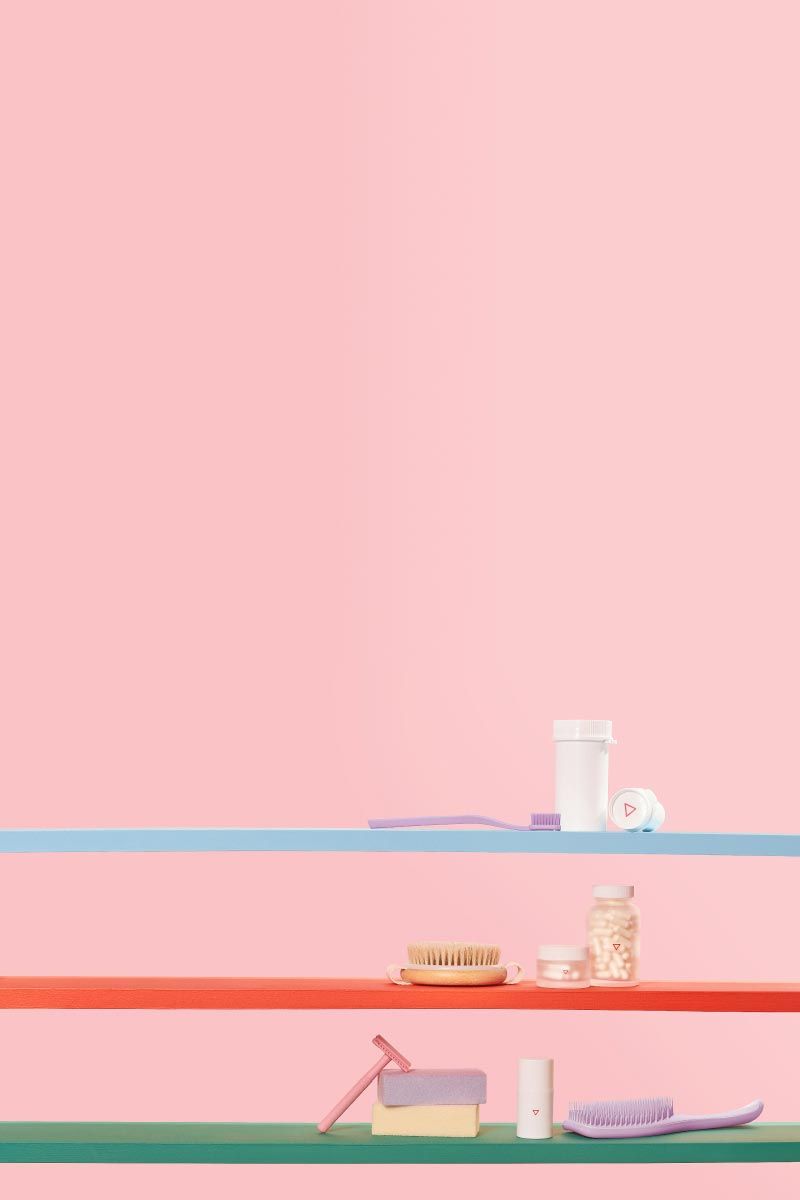 A variety of Wisp products on 3 colorful shelves with a purple toothbrush, a purple hairbrush, and two bars of soap in front of a pink background