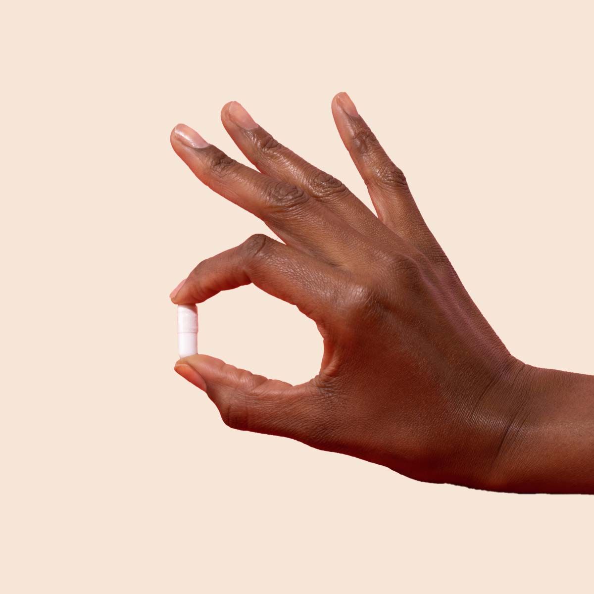 A person's hand holding a white capsule in front of a cream background