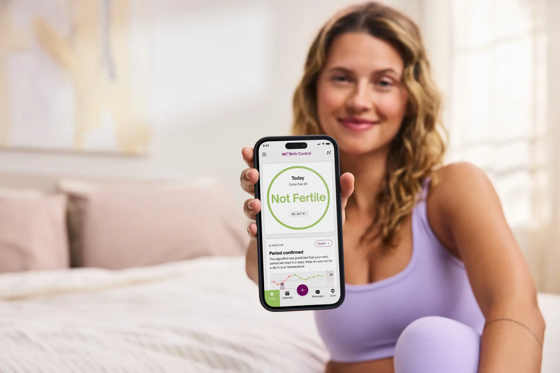 woman sitting on a bed in a purple workout clothing set holding a phone with the natural cycles app displayed