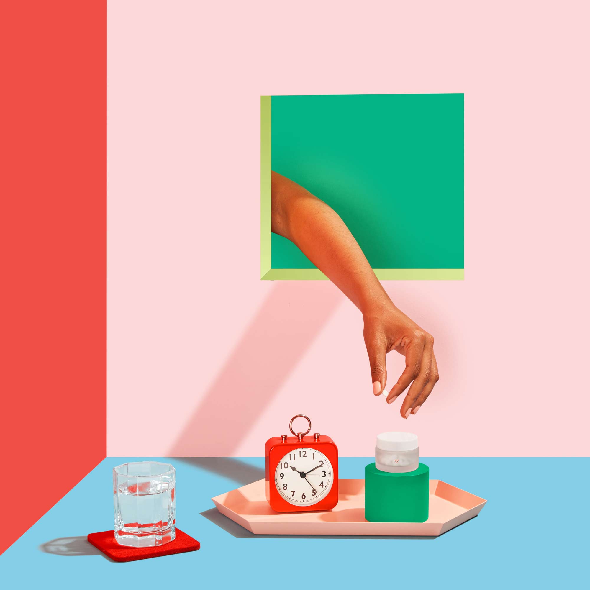 Hand reaching for Norethindrone pills next to an alarm clock, glass of water and colorful abstract shapes on a pink, red and blue background