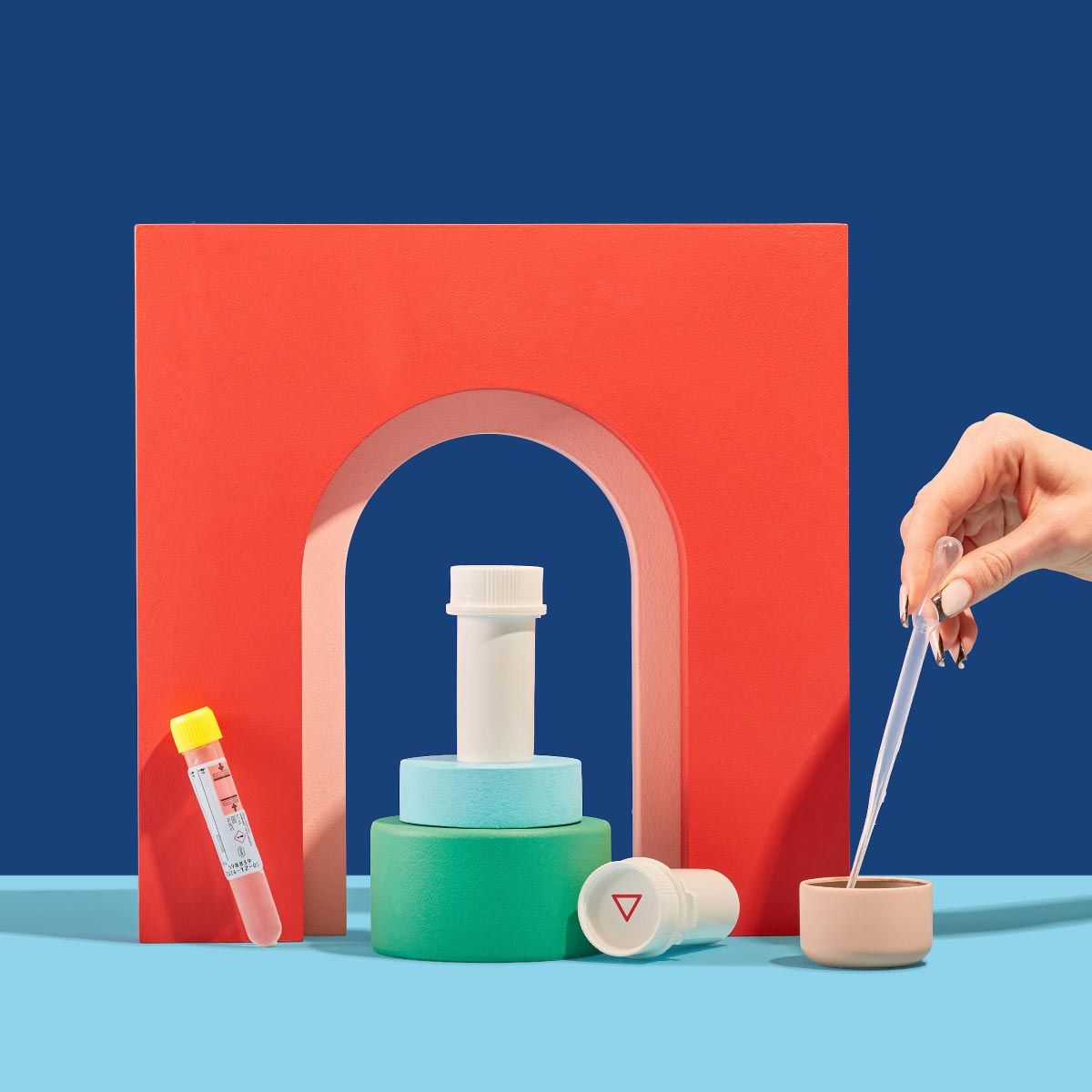 Wisp pill vials, testing tubes and a hand holding a testing pipette with colorful abstract shapes on a blue surface
