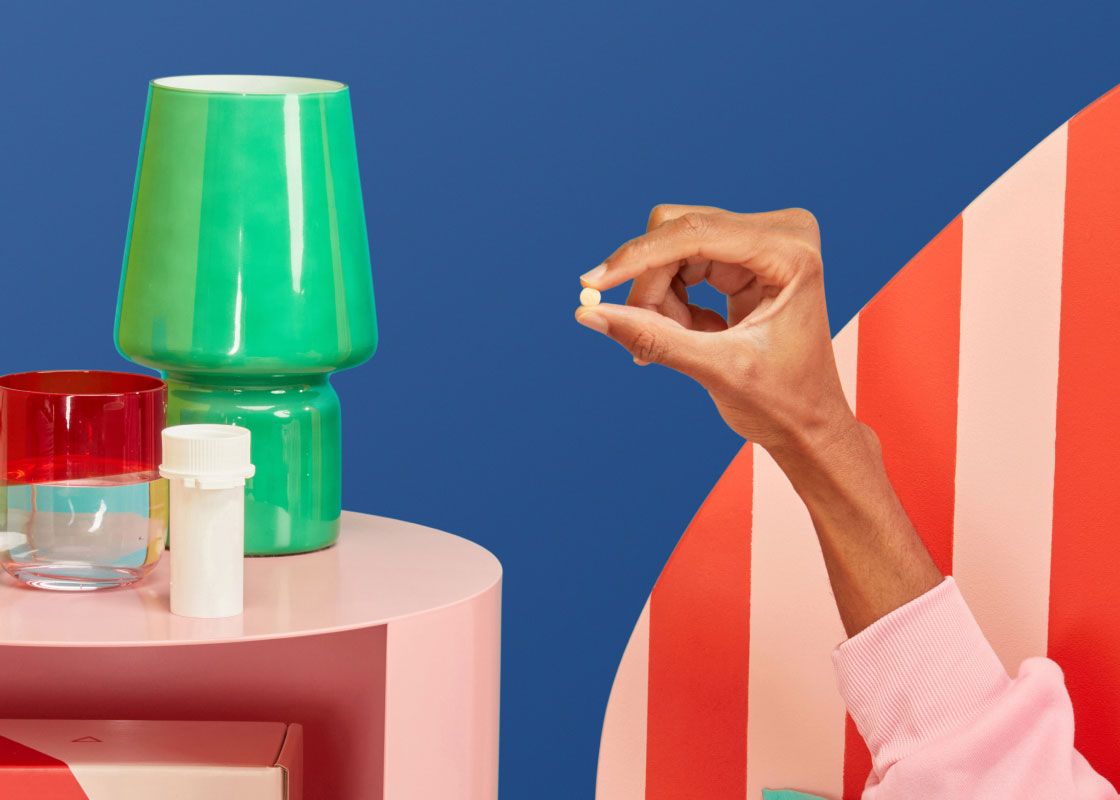 A man's hand holding a DoxyPEP pill next to a pink nightstand will a Wisp pill vial, a green lamp, and a glass of water