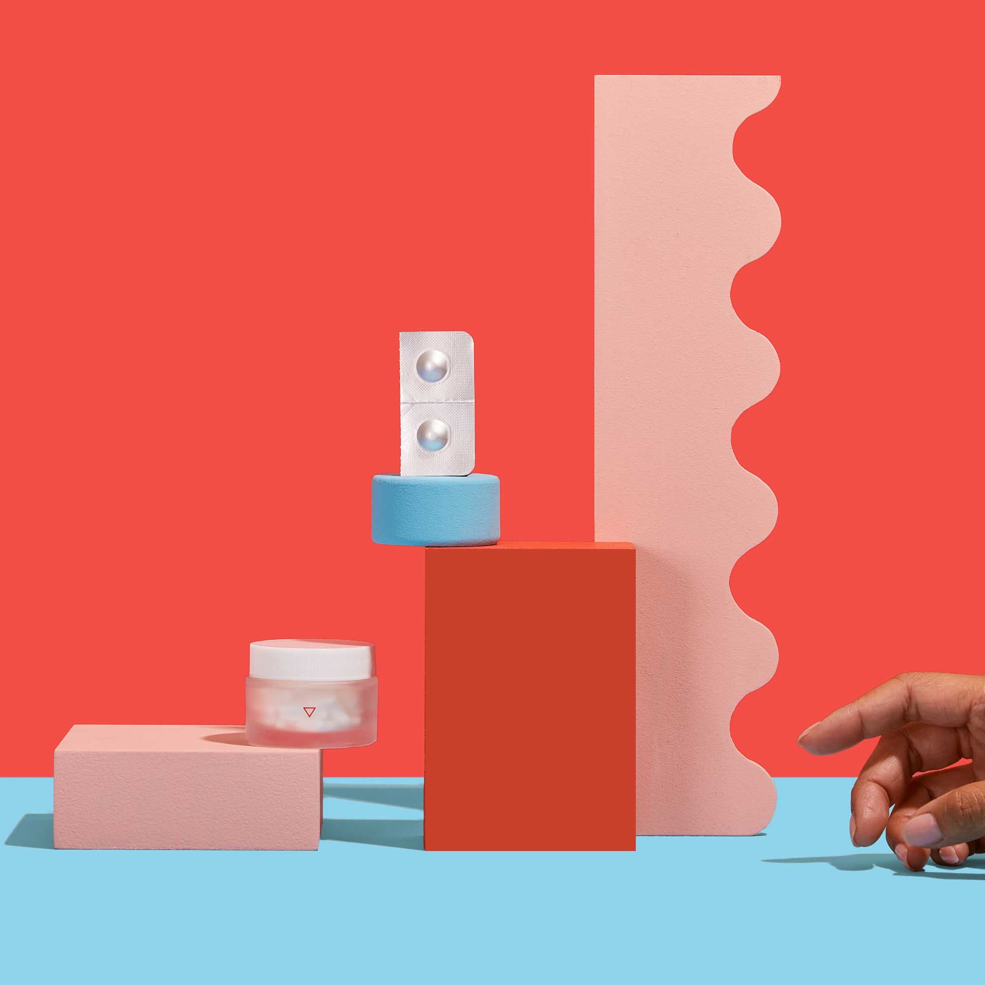 A Zofran pill packet and Wisp glass pill jar balanced on colorful abstract shapes on a light blue surface with a red background