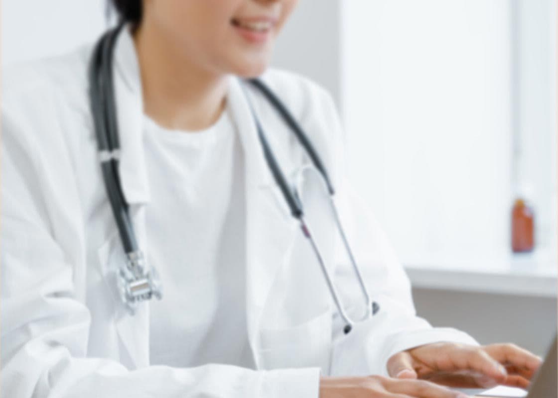 A female medical professional wearing a lab jacket and stethoscope is typing on a laptop