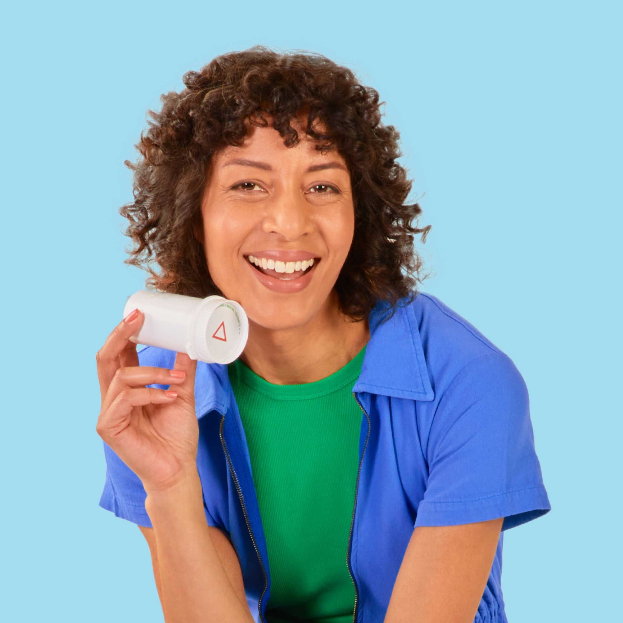 A woman with curly hair wearing a blue jumpsuit and a green shirt is smiling while holding a Wisp pill vial in front of a light blue background