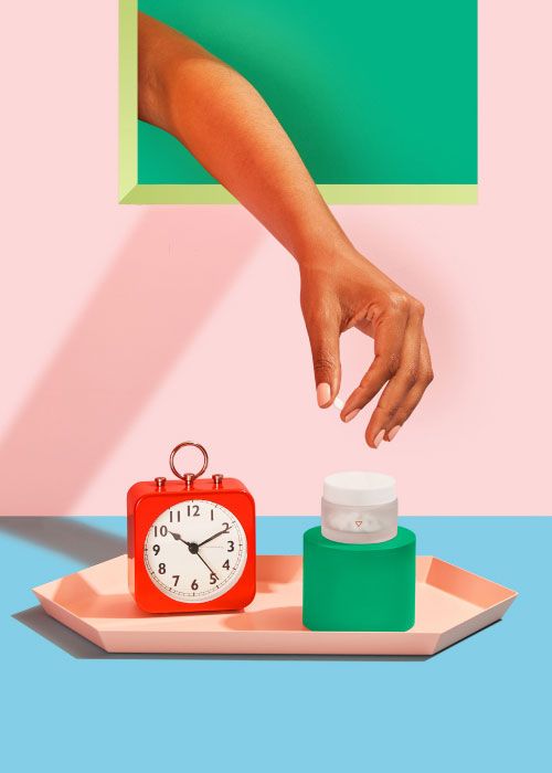 A woman's hand reaching for a small glass pill jar on a pink tray with a red alarm clock on a light blue surface with a pink background
