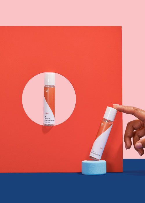 A woman's hand touching a bottle of Wisp Toy-Safe Lube and another bottle of lube balanced on colorful abstract shapes on a blue surface with a pink background