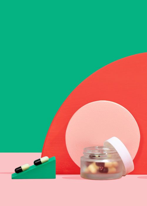 A small glass pill jar with two pills balanced on colorful abstract shapes on a pink surface with a green background