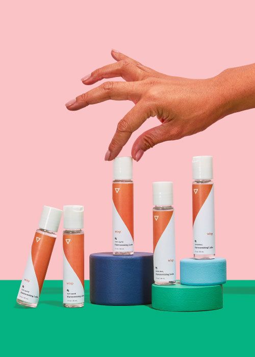 5 bottles of Wisp Harmonizing Lube balanced on colorful abstract shapes with a woman's hand reaching for one of them on a green surface with a pink background