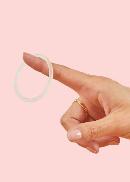 A woman's hand with NuvaRing  dangling off of her pointer finger in front of a pink background