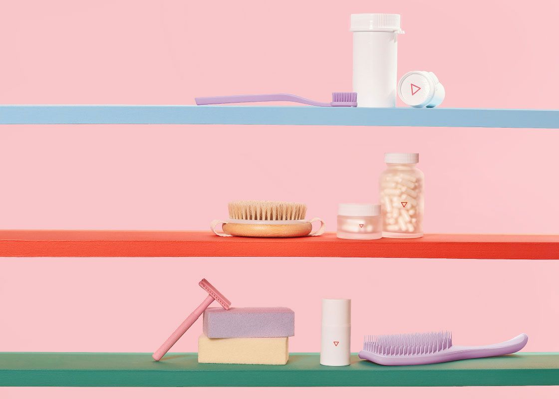 Various Wisp jars, vials, and bottles on colorful shelves with a purple brush, pink razor, purple toothbrush and colorful bars of soap in front of a pink background