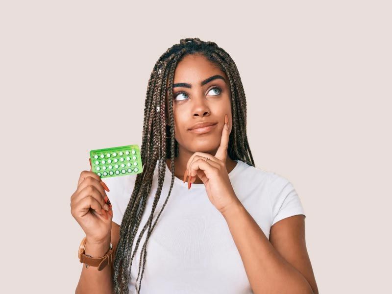 A woman wearing a white shirt and holding a green birth control packet with an expressing of contemplation on her face