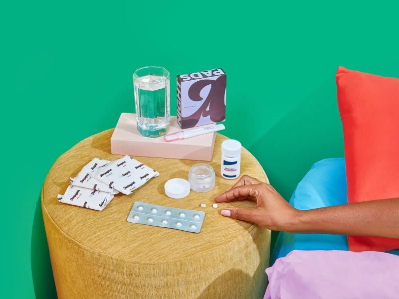 A woman's hand reaching for pills on a yellow nightstand with Wisp's medication abortion kit in front of a green wall with red, blue and pink bedding