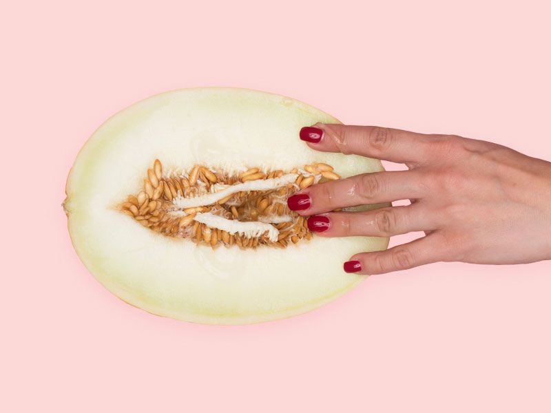 A woman's hand touching a vulva-shaped cut open fruit on a pink surface