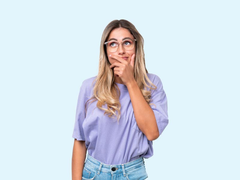 A woman with blonde hair wearing a purple shirt and jeans with a confused look on her face and a light blue background