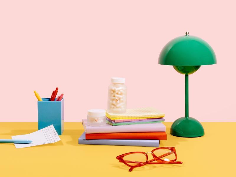 Wisp glass jars, a green lamp, pen holder, red glasses and an Rx script on a yellow and pink background