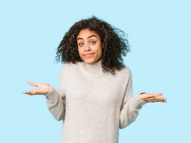 A woman wearing a grey sweater is shrugging in front of a light blue background