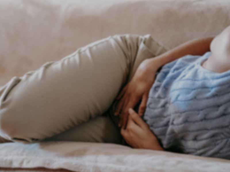 A woman wearing a blue sweater and tan pants is laying on a sofa with her hands on her abdomen