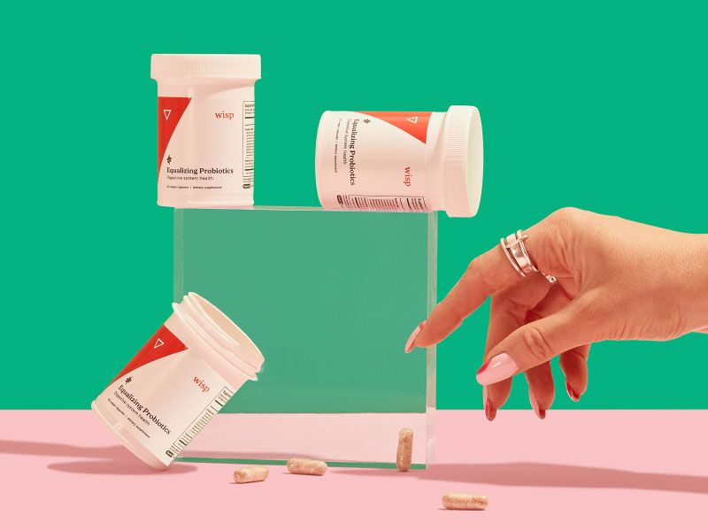 A woman's hand reaching for Wisp Equalizing Probiotics with 3 pill bottles balanced on clear abstract shapes on a pink surface with a green background