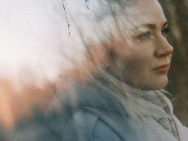 A woman gazing thoughtfully into the distance, with a soft double-exposure effect blending her face with a blurred natural background.