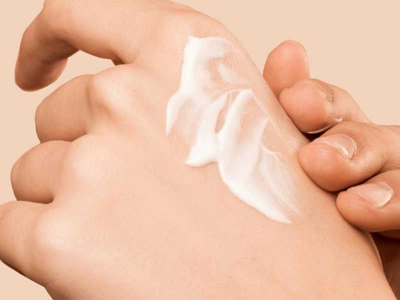 Hands applying white moisturizer or lotion against a peach background.