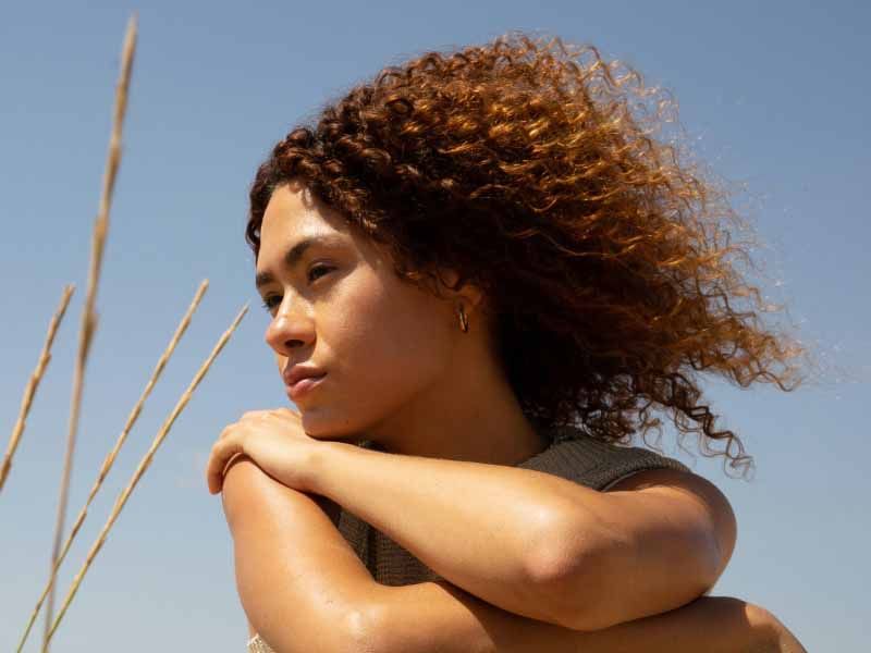Woman with curly hair looking into the distance outdoors, with tall grass and a blue sky behind her.