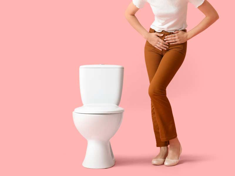 A woman standing next to a toilet wearing a white shirt and brown pants with her hands on her pelvic region indicating discomfort