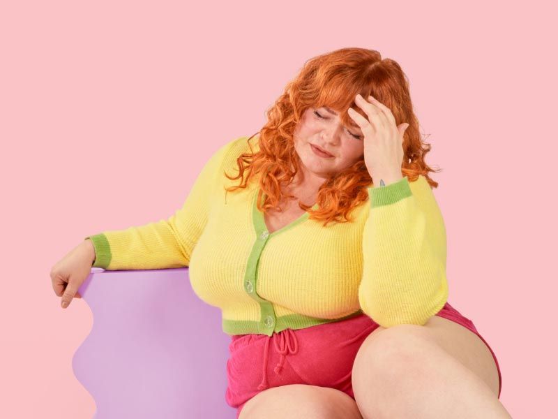 A woman wearing a yellow and green sweater and red shirts has her hand on her forehead in distress