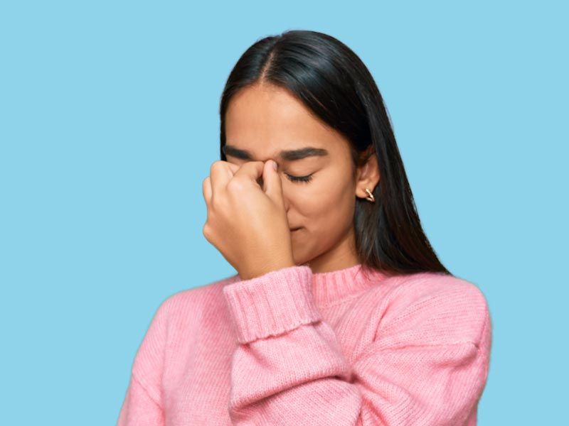 A woman with dark hair and a pink sweater is pinching the bridge of her nose in frustration