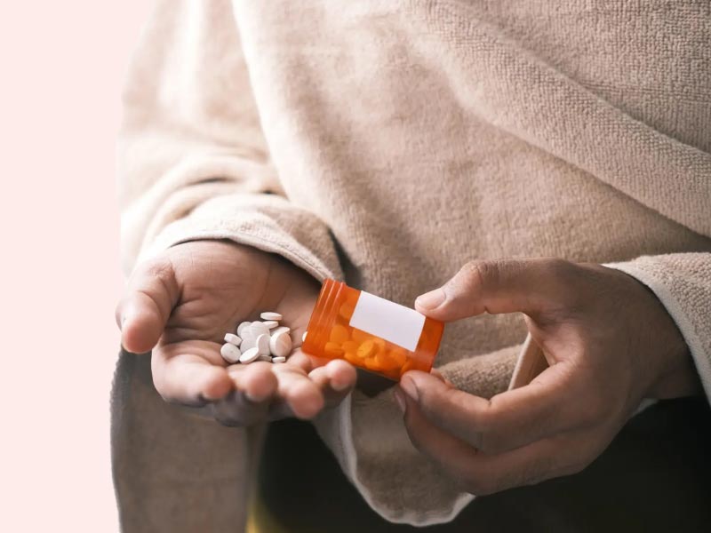 A woman wearing a tan shawl and brown pants holding a pill bottle and dumping small white pills into her hand