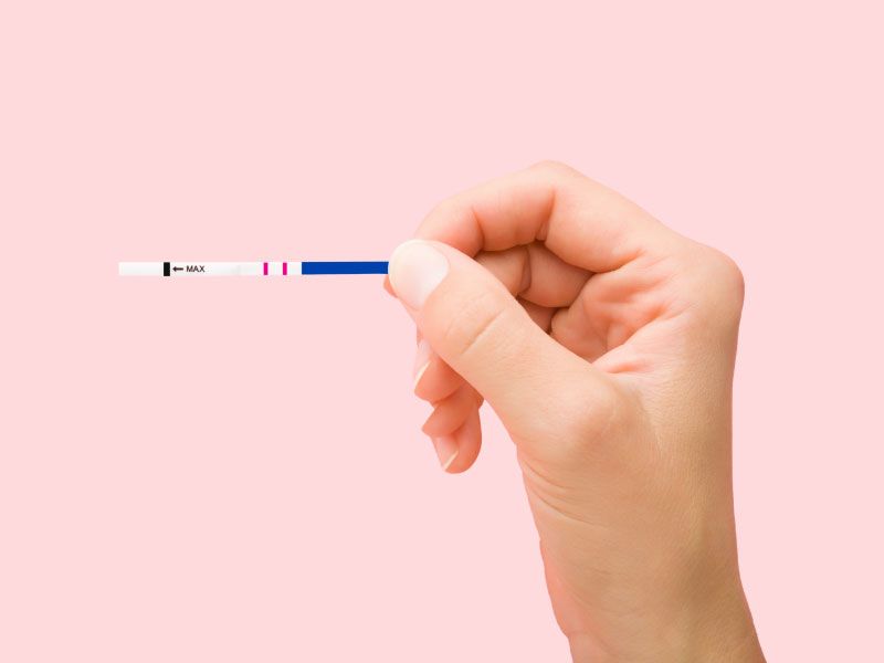 A person's hand holding a testing strip in front of a pink background