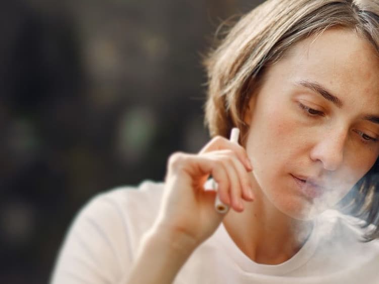 A woman in a white T-shirt sits holding a vape pen, exhaling a cloud of vapor as she looks downward.