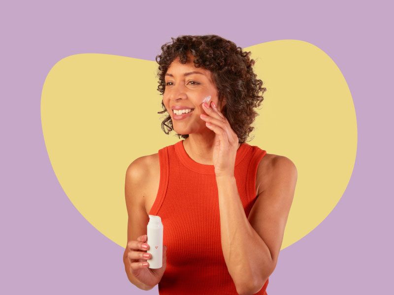 A woman wearing a red tank top is holding a bottle of Estriol cream and applying it to her face