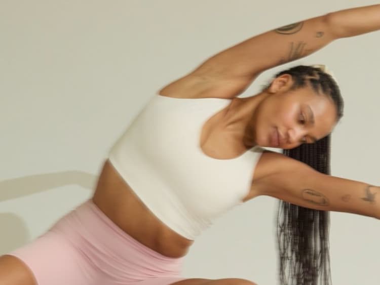 A woman in a cream crop top and pink shorts does a standing side stretch against a plain wall.