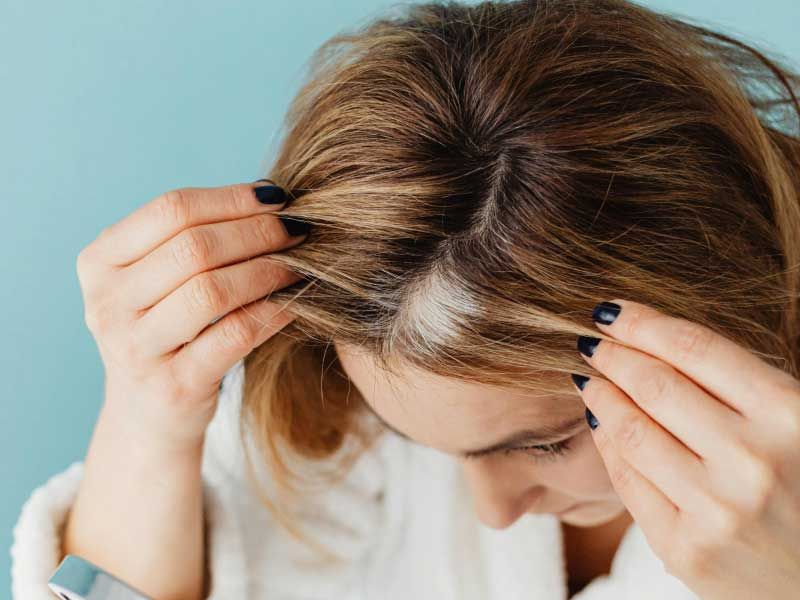 A woman wearing a white robe is looking at her thinning hair in the mirror with a teal background