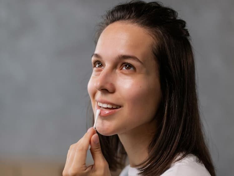 A smiling woman wearing a white shirt is applying a cream to her lip with a q tip
